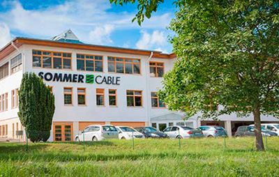 A picture of our company building in Straubenhardt under a blue sky. A green meadow and parked cars can be seen in front.