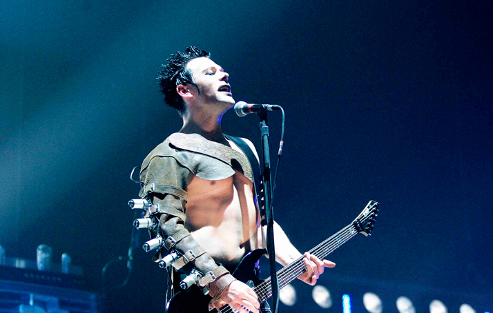 A photo of the artist Richard Z. Kruspe during a performance. He holds his guitar in his hands and sings into a microphone.