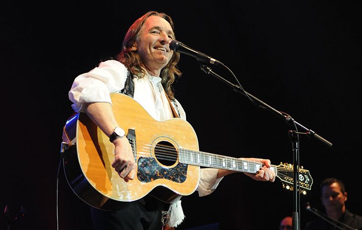 A picture of our artist Roger Hodgson on stage. He holds a guitar in his arms and smiles into the microphone in front of him.