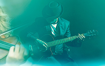 A musician in the middle playing the guitar. On the left side is a detail of a person playing the violin. The picture has a turquoise veil.