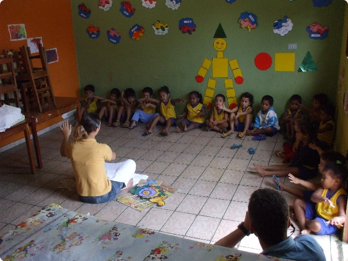 Mehrere Kinder sitzen in einer Reihe auf dem Boden. Vor ihnen sitzt eine Frau und hebt ihre linke Hand.