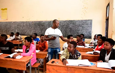 A picture of our school and education project eQUiP-Magu in Rwanda. The picture shows a school class. The teacher is standing in the middle and the pupils are sitting at their desks. 