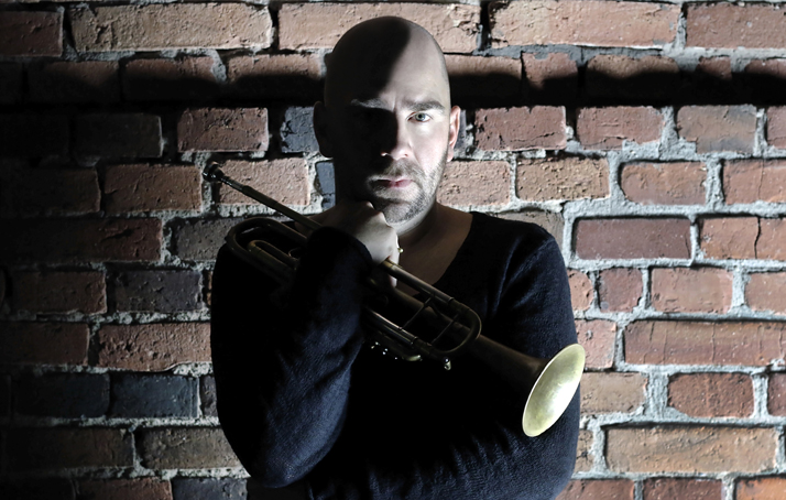 A photo of the artist Sebastian Studnitzky in front of a stone wall. He is holding a trumpet in one hand.